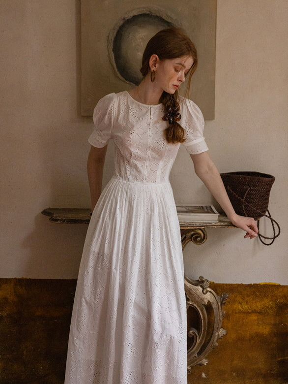 A woman standing by a vintage console, wearing a white eyelet lace dress with puff sleeves and a braided hairstyle. Timeless cottagecore style.