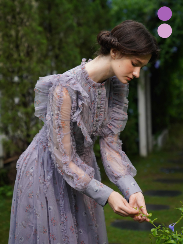 A woman in a lavender floral ruffle dress gently touches a flower, capturing a quiet fairycore moment in a lush garden.