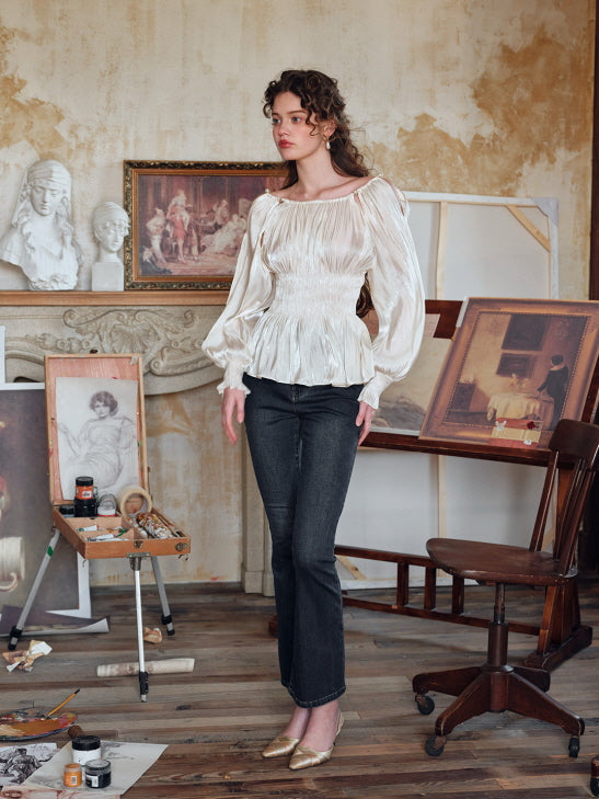 Fairycore-inspired ivory satin blouse paired with black jeans, featuring long puff sleeves and a shirred waist in a classic atelier backdrop with vintage artwork.