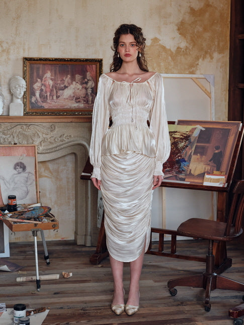 A woman wearing an ivory satin blouse with puff sleeves and a ruched waist, paired with a flowing cream skirt, standing in an art studio filled with vintage drawings and classical painting tools.