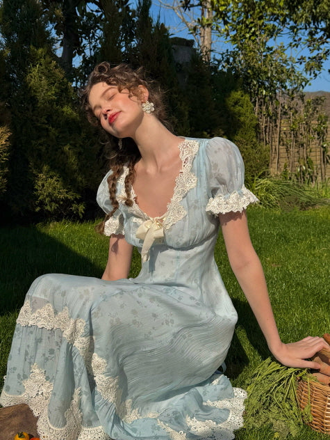 A woman sitting gracefully on the grass in a romantic light blue lace-trimmed dress, with her eyes closed and head tilted toward the sun, surrounded by greenery and rustic charm.