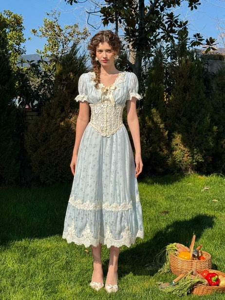 A woman wearing a light blue romantic dress with a decorative ivory lace corset, puff sleeves, and scalloped hemline, standing on grass beside a picnic basket filled with fresh vegetables.