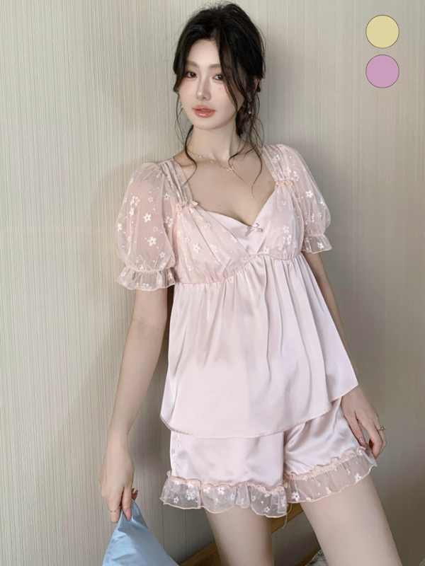 Model leaning against a wall in a blush pink fairycore pajama set with ruffled hems and embroidered sheer sleeves, holding a pale blue cushion.