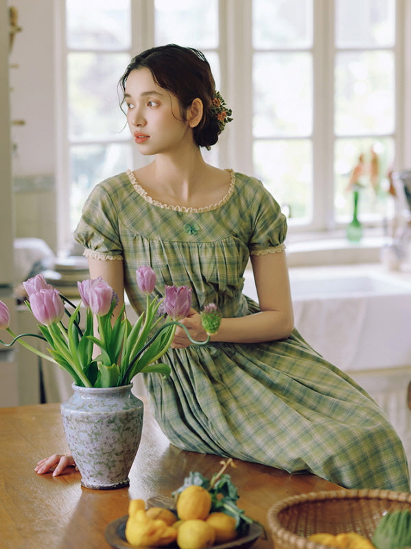A serene moment as the model sits atop a wooden table, surrounded by fresh tulips and lemons, wearing a green plaid dress adorned with delicate lace trim and a small floral accent.
