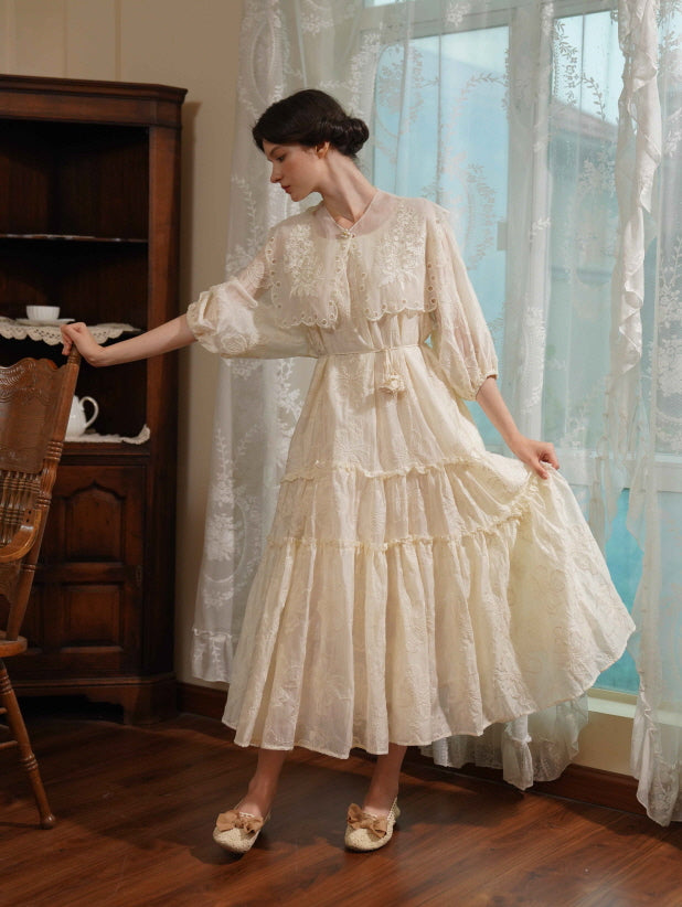A woman in a vintage-inspired cream tiered dress poses gracefully by a wooden chair, evoking a soft romantic cottagecore mood.