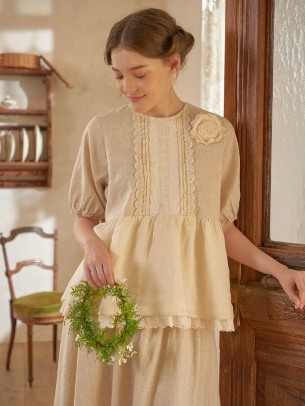 A young woman in a vintage-inspired beige blouse and skirt stands by a wooden door, holding a small floral wreath in a cottagecore setting.