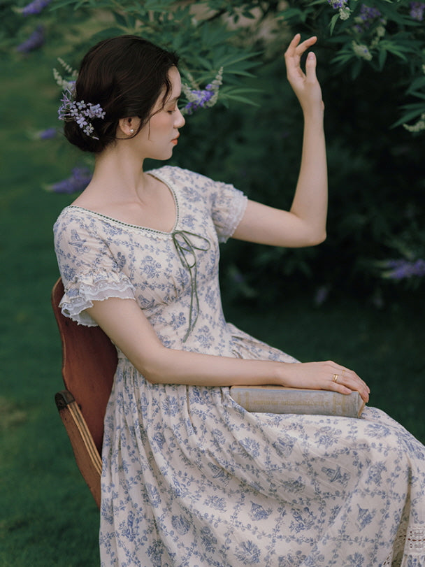 Seated amidst lush greenery, the model wears a soft floral dress with lace sleeves and a delicate ribbon tie, creating a serene, fairytale-like scene.
