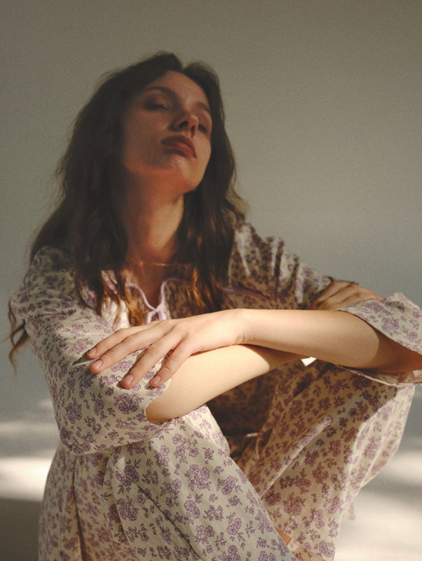 The model sits in soft sunlight wearing a vintage-inspired lavender floral loungewear set, capturing a dreamy and peaceful cottagecore mood.