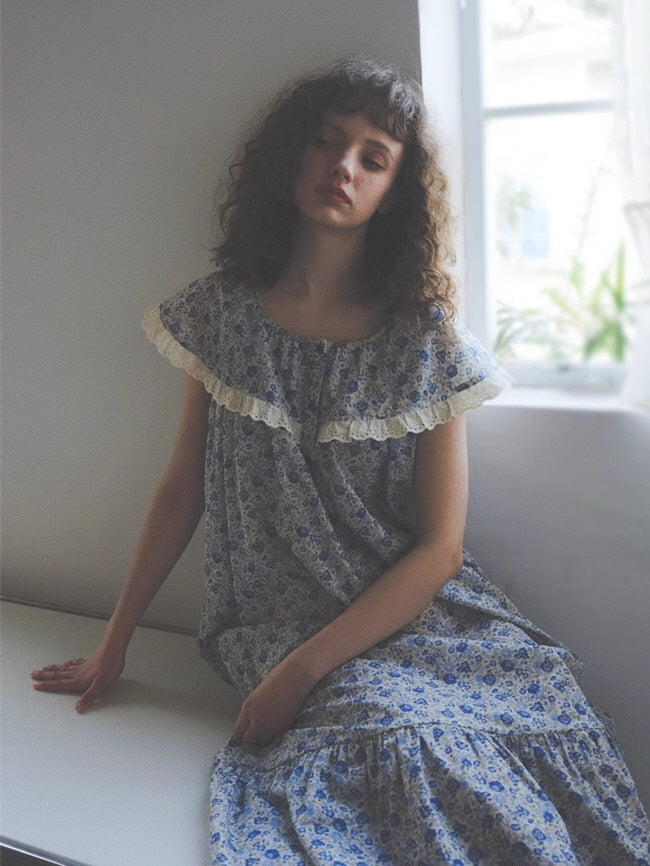 A young woman in a vintage-style blue floral nightgown with lace-trimmed cape collar sits dreamily by a window, evoking a cottagecore mood.