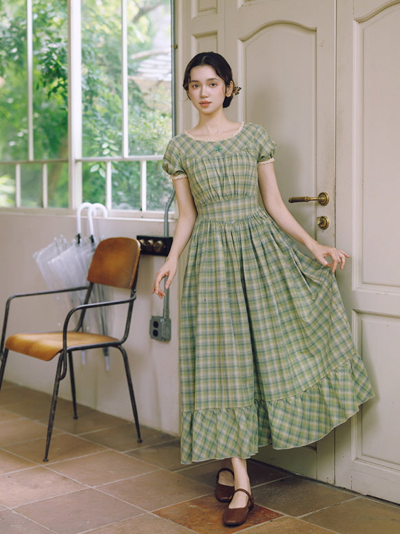 A young woman in a vintage-inspired green plaid dress, standing gracefully in a cozy, sunlit room. The dress features a soft, cottagecore aesthetic with puffed sleeves and a flowing silhouette.