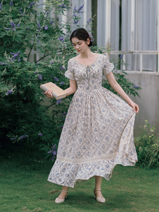 A romantic cottagecore dress with blue floral prints and lace trim, as the model holds a book and gently lifts the hem, evoking a vintage-inspired moment.