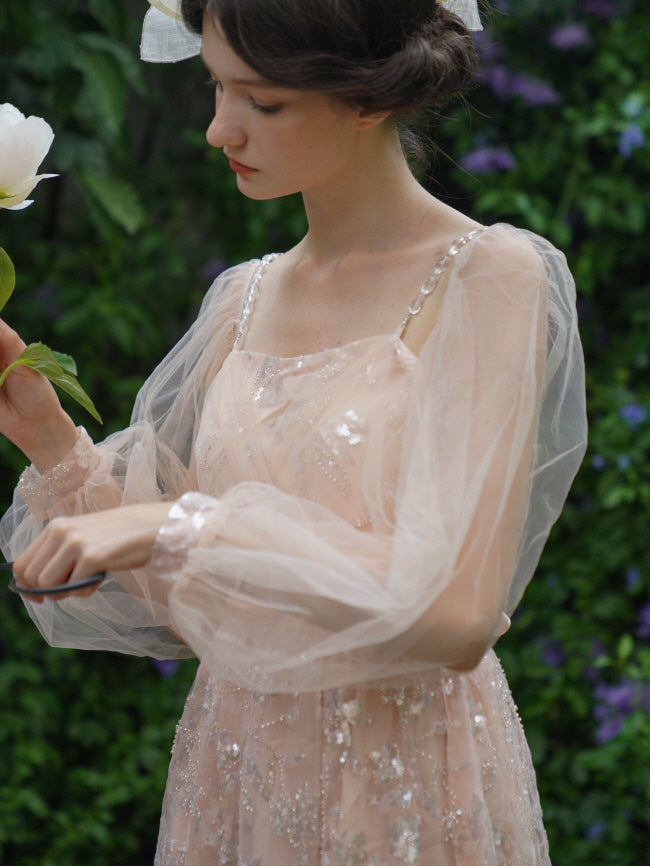 Model gazes at a flower while wearing a blush pink dress with glittering embroidery and sheer puff sleeves, evoking a dreamy fairycore mood.