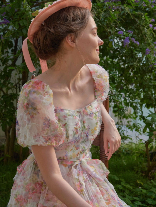 A woman in a puff-sleeve floral dress with a square neckline and ribbon tie sits gracefully in a garden, styled in a dreamy fairycore look.