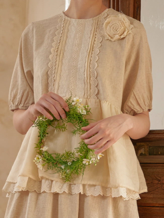 Close-up of a woman wearing a cottagecore blouse with lace panels and a fabric rose, gently holding a flower wreath in a warm, romantic light.