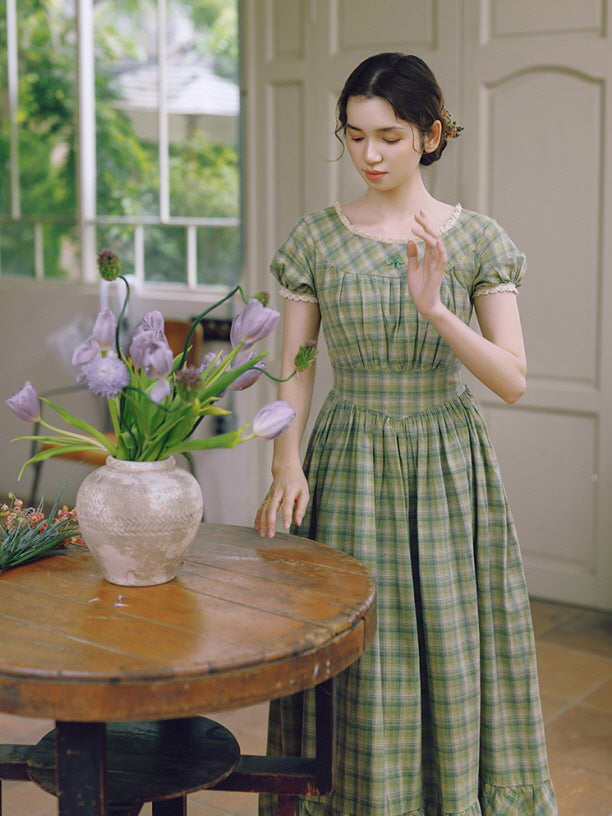 A young woman in a vintage-inspired green plaid dress gently touches lavender tulips on a wooden table. The soft, cottagecore style dress features a delicate lace neckline and puff sleeves, creating a dreamy, fairytale ambiance.