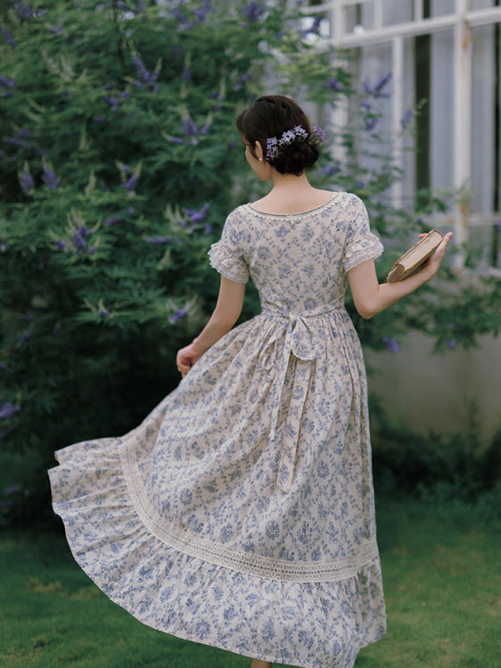 Back view of a flowy, vintage-inspired floral dress with lace trim and a ribbon tie, as the model gracefully walks holding a book, evoking a fairytale charm.