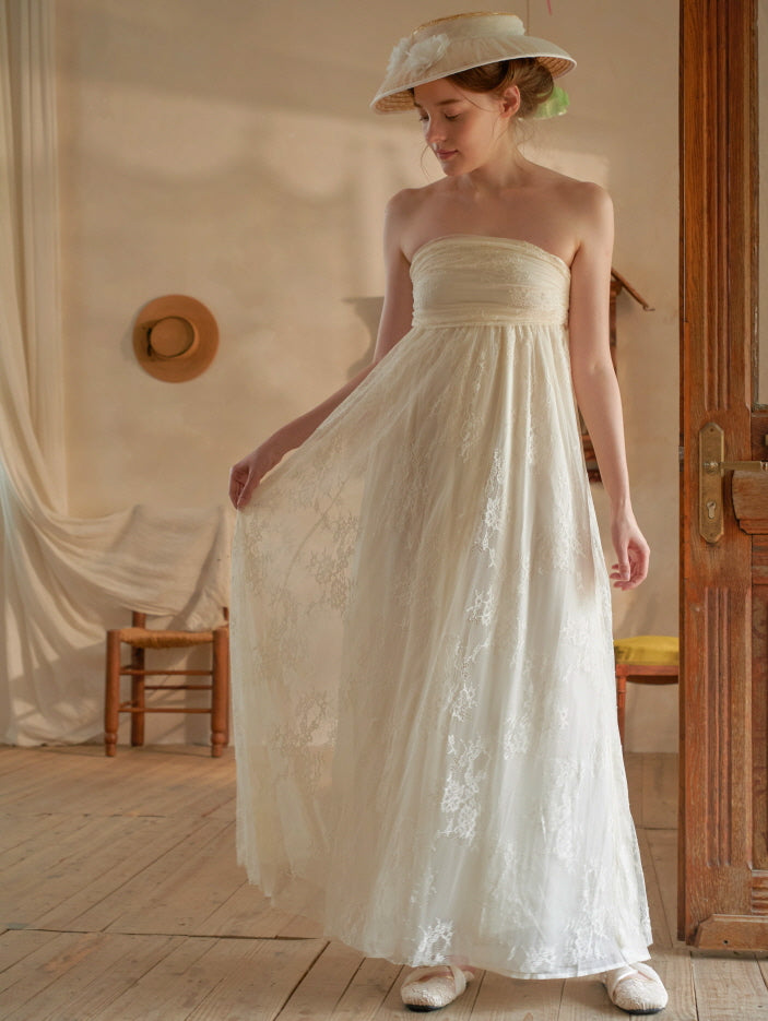 Wearing a lace-embroidered strapless ivory dress, the model stands gracefully in a cozy vintage room filled with fairytale charm.