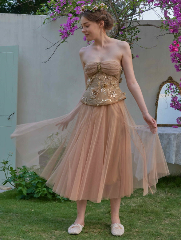 A woman twirls gracefully in a nude-toned strapless tulle dress with floral embroidery, radiating a dreamy fairycore garden look.