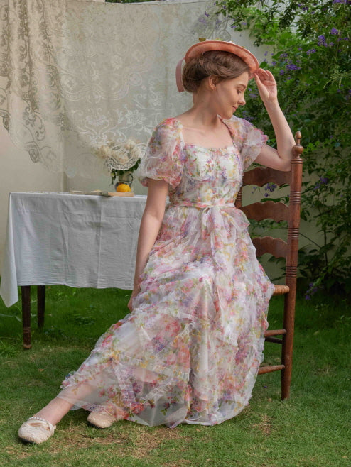 Seated in a garden, the woman tilts her bonnet gently while wearing a pastel floral puff-sleeve dress with ruffled layers and fairycore charm.