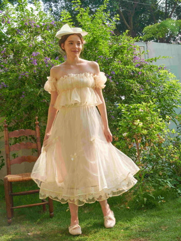 The model smiles in a soft ivory tulle skirt with lace and ribbon bows, styled in a fairycore garden setting with a vintage-inspired top and hat.