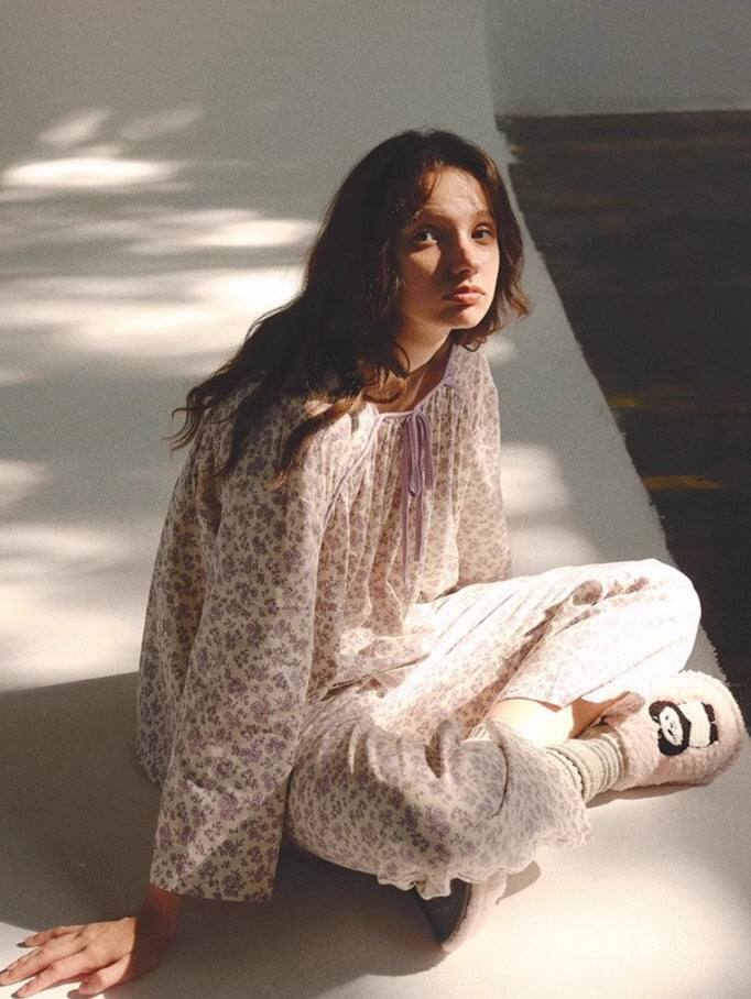 Wearing a relaxed lavender flower print pajama set, the model sits on the floor in warm light, evoking a cozy fairytale loungewear aesthetic.