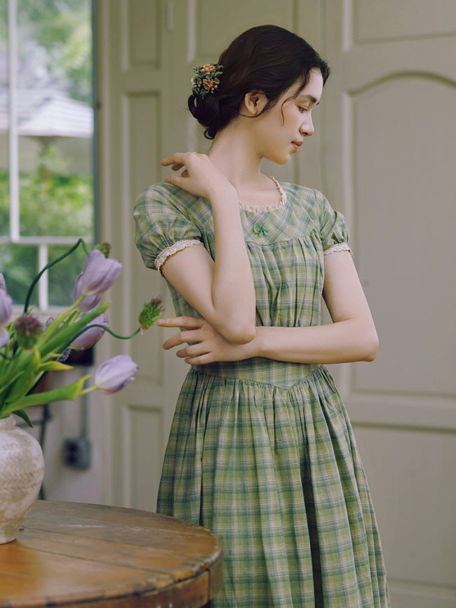 A serene moment as the model stands gracefully beside a wooden table adorned with lavender tulips, wearing a vintage-inspired green plaid dress with delicate lace detailing and a soft, cottagecore aesthetic.