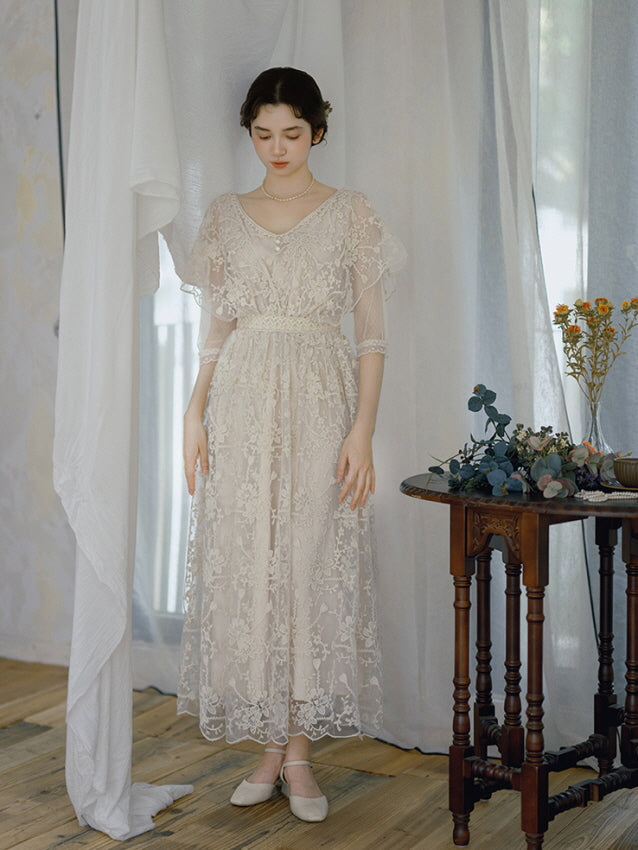 Wearing a soft ivory lace dress with flutter sleeves, a woman stands gracefully by a wooden table and white curtains, evoking a fairytale mood.