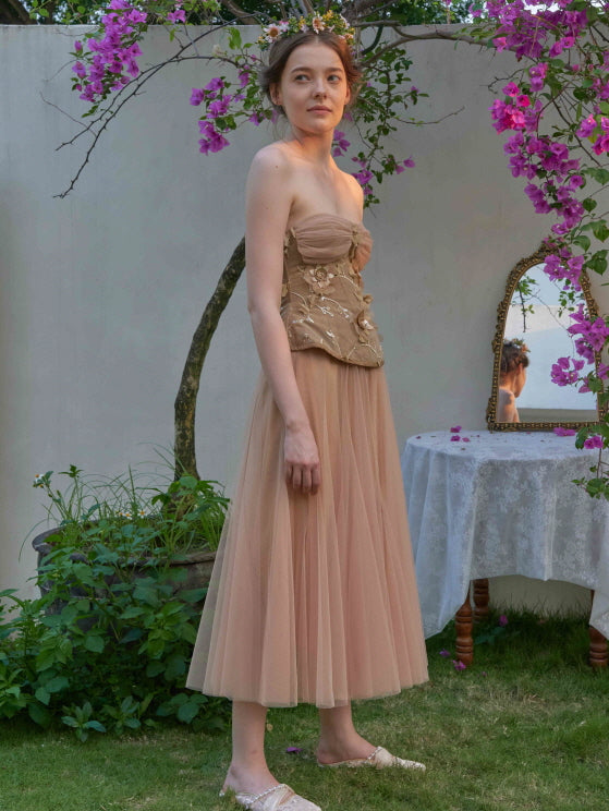 A woman in a nude strapless floral dress stands in a blooming garden, styled with embroidered details and a soft princesscore silhouette.