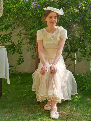 Seated gracefully in a garden, the model wears a delicate lace-trimmed tulle skirt paired with a soft embroidered top, creating a gentle fairycore look.