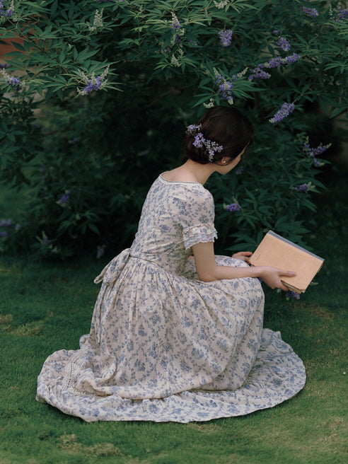 A serene, vintage-inspired moment as the model kneels on the grass, holding a book while wearing a delicate blue floral dress with lace accents and a bow-tied back.