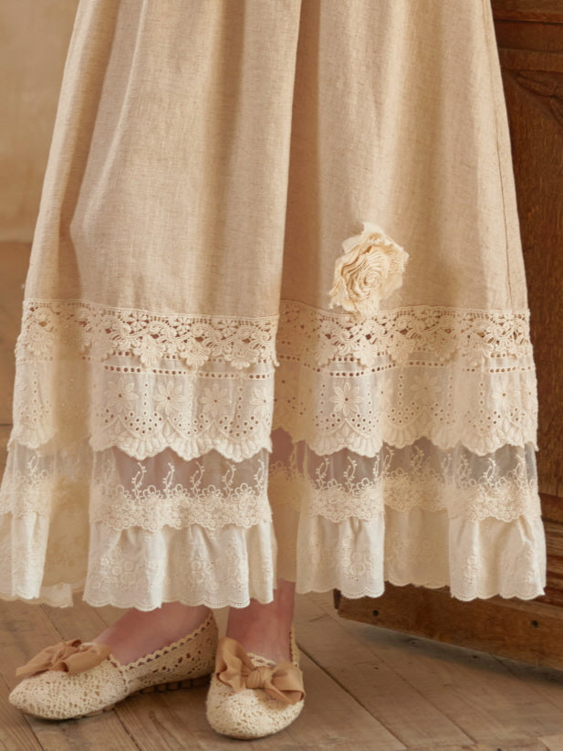 Close-up of a beige cottagecore skirt with layered lace hem and fabric flower detail, paired with crochet bow shoes on wooden flooring.