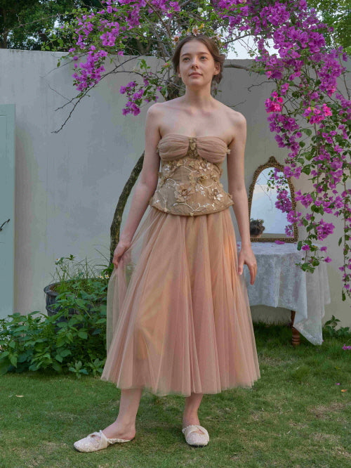 Wearing a nude tulle dress with floral embroidery, the model stands under blooming trees in a dreamy garden, evoking a soft princesscore mood.