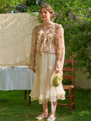 Wearing a floral chiffon blouse with a ribbon tie and a layered ivory skirt, the model evokes a vintage cottagecore charm in a lush outdoor scene.