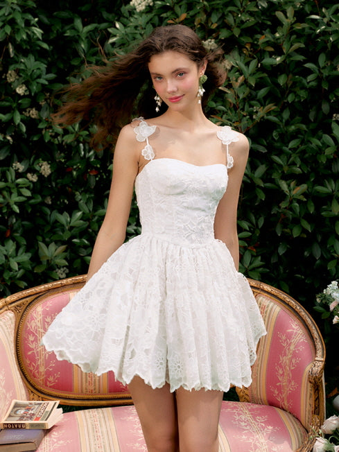 A young woman stands with wind-blown hair in a white lace mini dress, radiating a fairycore charm in a dreamy garden setting.