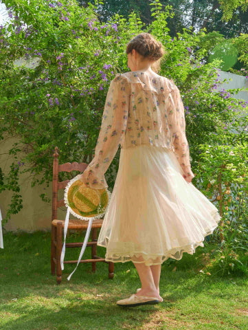 Back view of a flowing tulle skirt paired with a floral blouse, as the model holds a straw bonnet in a dreamy princesscore-inspired outdoor scene.