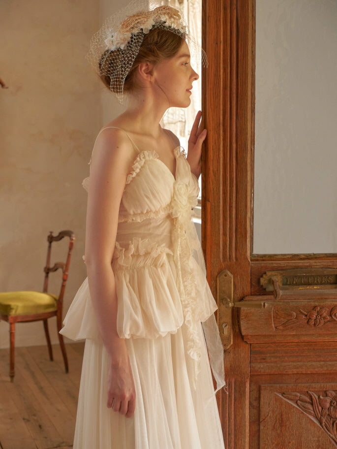 A side view of a model in a vintage-inspired cream dress with gathered ruffles, standing near a wooden door in soft morning light.