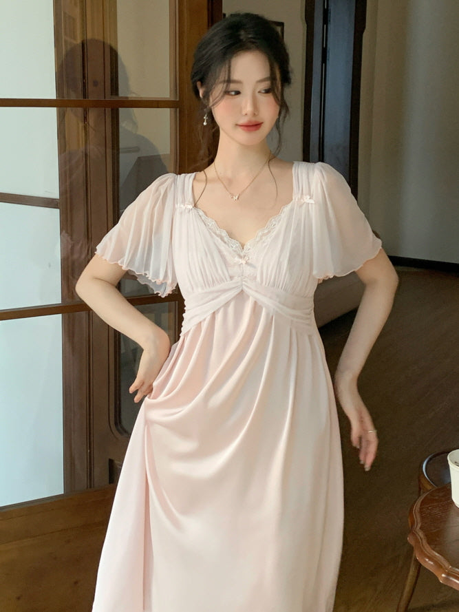 A woman in a soft pink fairycore nightgown with flutter sleeves and lace trim stands gracefully in a wooden interior, evoking a romantic mood.