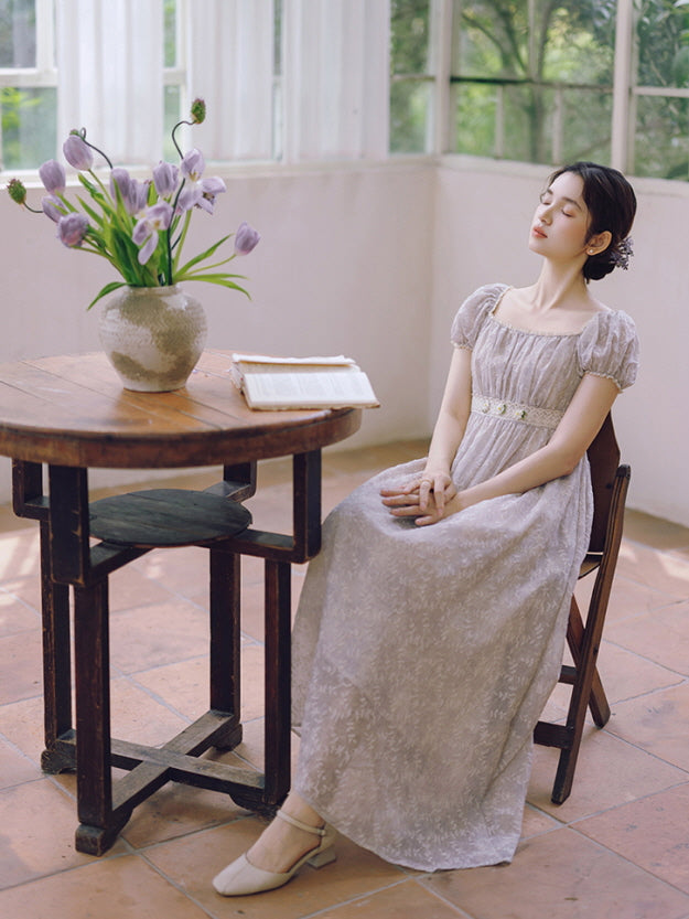 A woman in a lavender vintage dress with floral embroidery sits peacefully by a wooden table, surrounded by soft morning light and tulips.