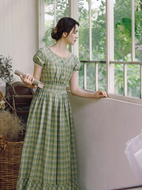 A serene moment as the model gazes outside, wearing a vintage-inspired green plaid dress with puff sleeves and lace trim. Cottagecore aesthetic.