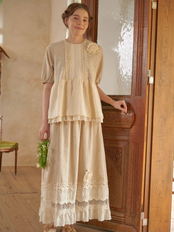 A woman stands in a warm-lit room wearing a cottagecore outfit with puff sleeves, lace trim, and a soft beige floral skirt, holding a wreath.