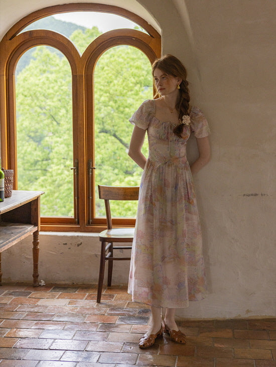 A young woman wearing a pastel watercolor floral dress with puff sleeves stands by an arched window, exuding a dreamy, vintage cottagecore vibe.