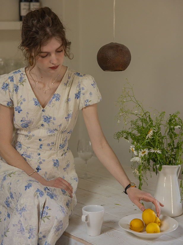 A romantic floral midi dress with blue accents, worn by a model seated at a rustic table, surrounded by white daisies and fresh lemons.