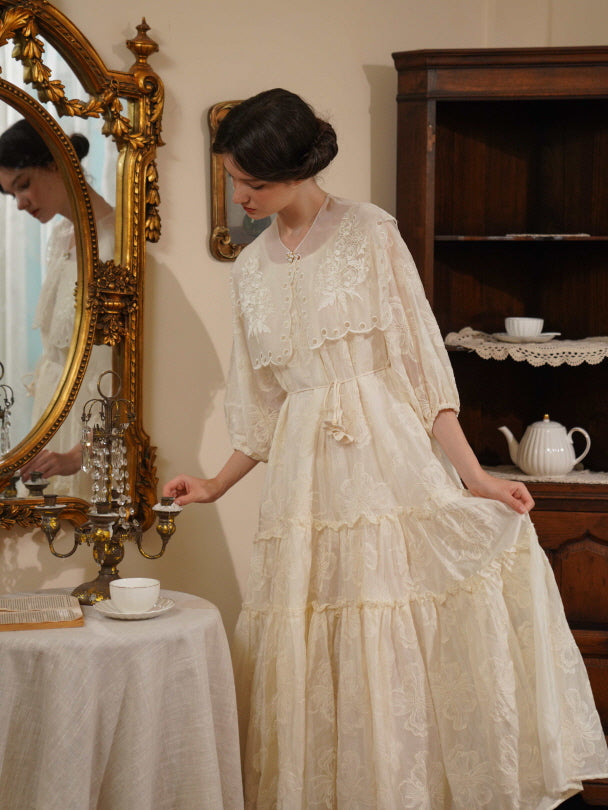 A woman in a tiered floral lace dress gently lifts her skirt while standing beside a golden mirror, capturing a timeless princesscore mood.