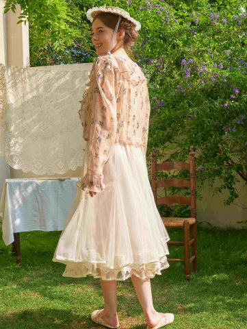 The model smiles over her shoulder in a sheer floral blouse and lace-trimmed ivory skirt, capturing a fairycore garden moment with vintage charm.
