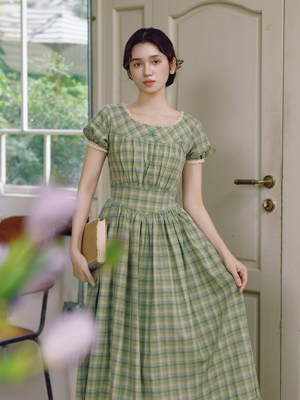 The model stands gracefully by a door, holding a book while showcasing the classic silhouette of a green plaid dress adorned with delicate lace and a waist tie. Vintage-inspired charm.