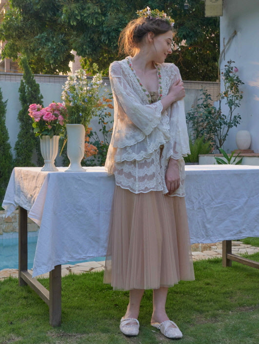 A woman stands by a garden table in a layered lace robe and tulle skirt, styled in a soft fairycore look with floral crown and slippers.