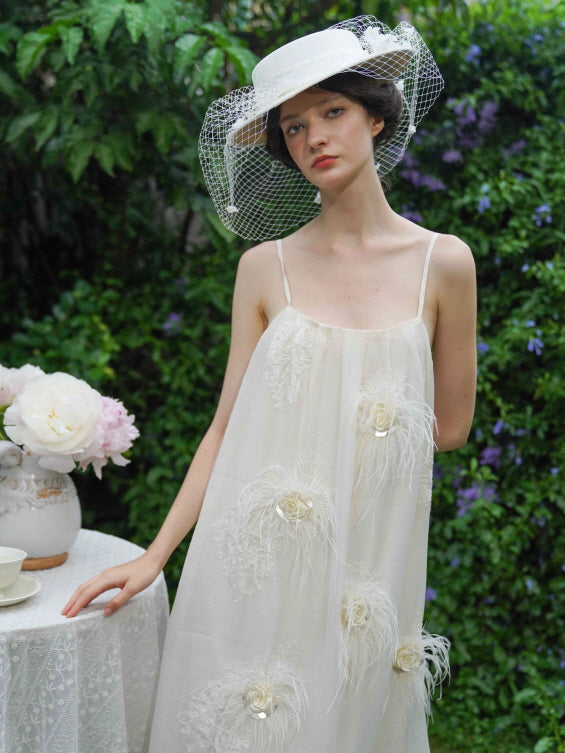 Model leans softly against a lace table, wearing a cream floral feather dress and vintage hat, capturing a serene fairycore garden moment.