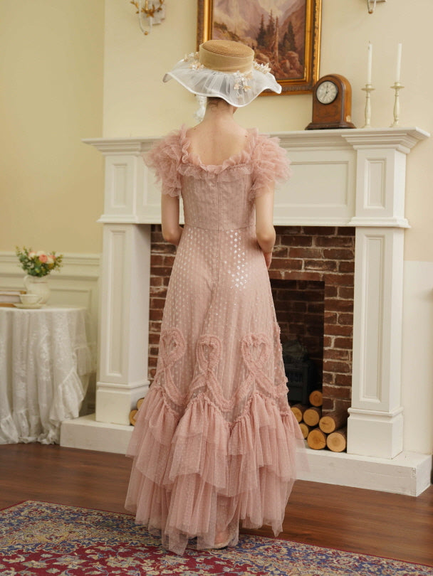 A woman in a vintage-inspired blush pink princesscore dress with ruffled layers and embroidered heart motifs poses in front of a white fireplace.