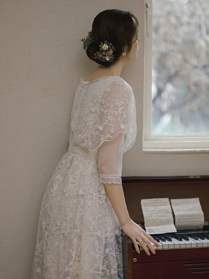 A woman in a romantic lace gown with floral hairpieces gazes out a window beside an antique piano, evoking vintage and cottagecore elegance.