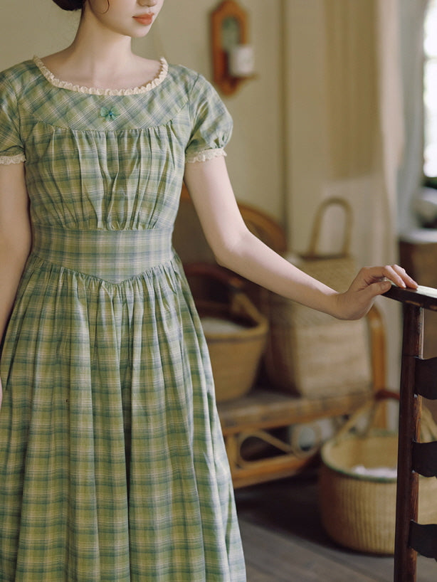 A close-up of a woman seated on a wicker chair, wearing a green plaid vintage-inspired dress with delicate lace trim along the neckline and puff sleeves. The soft, cottagecore aesthetic evokes a romantic, timeless vibe.